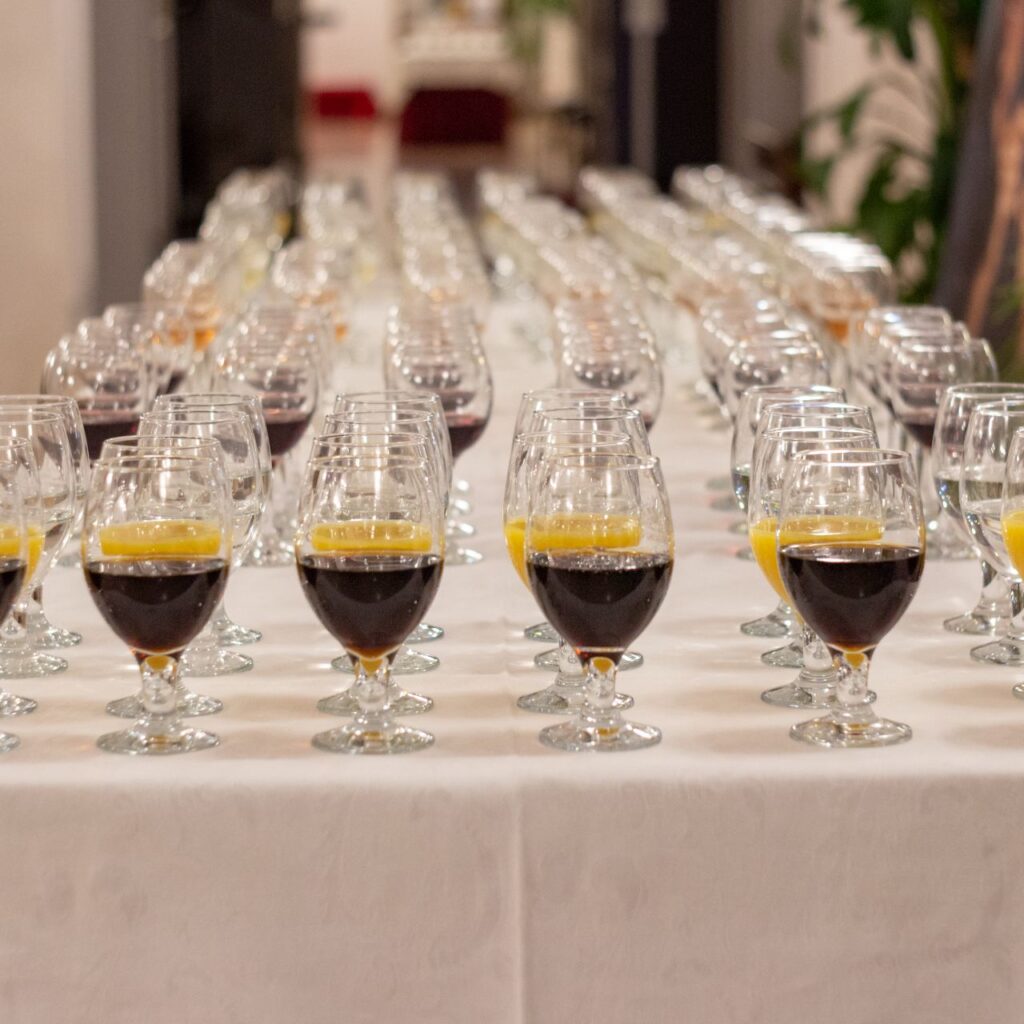 Egy asztal van a képen, amin fehér asztalterítő van. Az asztalon rengeteg sorba rendezett üvegpohár van, amikben üdítő és bor van