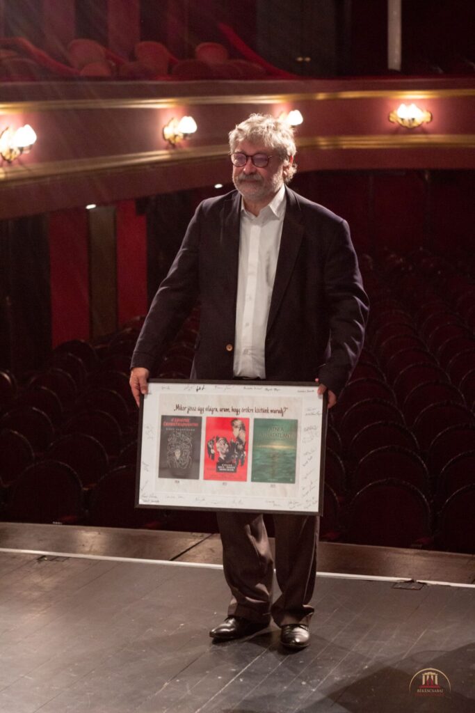 Seregi Zoltán igazgató úr áll a színpadon, mögötte a nézőtér. A kezében egy képkeretet tart, amiben három plakát látható