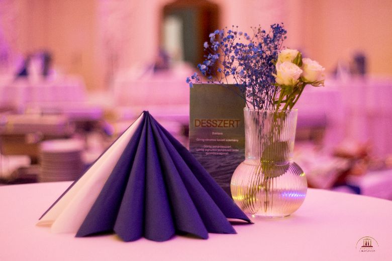 A képen egy asztalnak a teteje látható, amin bal oldalt fehér és kék szalvéták vannak szépen felállítva egy körben, mögötte egy étlap, jobbról pedig egy átlátszó üveg vázában kék kicsi virág és fehér rózsa van