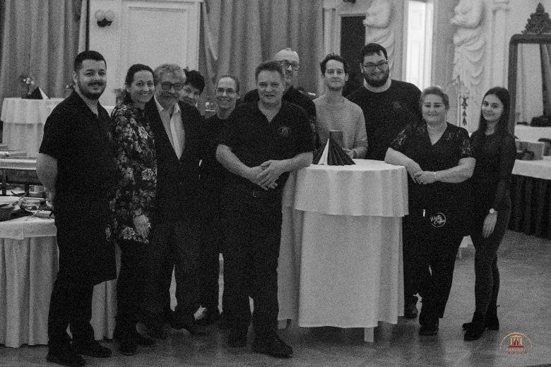 Pincérek állnak a képen a kamerába nézve mosolyognak, előttük egy fehér terítős kicsi kör asztal van, amin szalvéták vannak