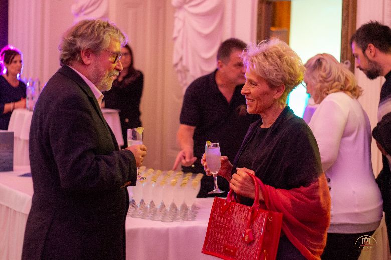 Egy öltönyös férfi áll a kép bal oldalán, kezében pezsgős pohár, jobb oldalt pedig egy idős hölgy áll szintén pohárral a kezében, és egymásra nevetnek