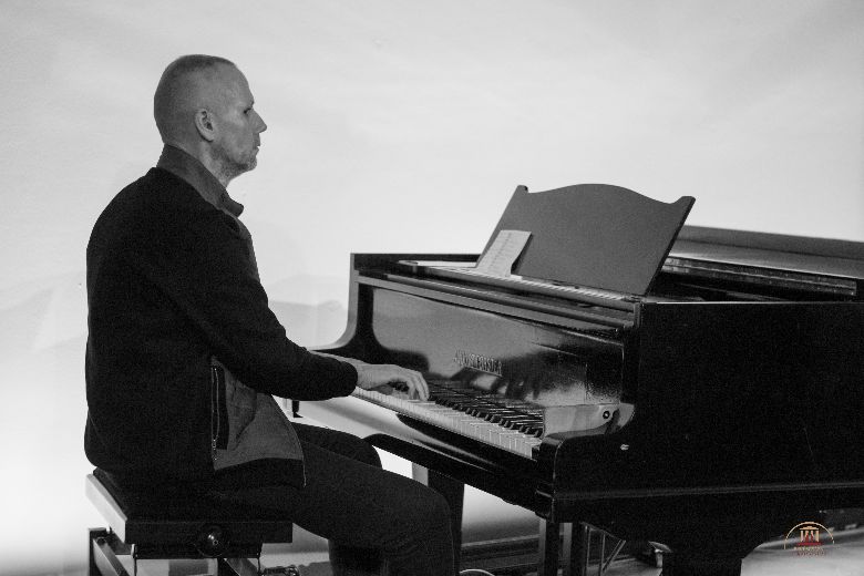 Bal oldalt egy férfi ül egy zongoraszéken, jobbról a zongora látható és a férfi éppen a zongorán játszik