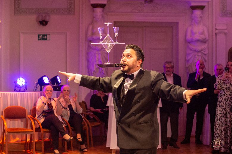 A képen egy öltönyt viselő férfi áll, a kezeivel maga mellett kinyújtva egyensúlyozik, a szájával egy tálcát tart, amin négy pezsgőspohár van