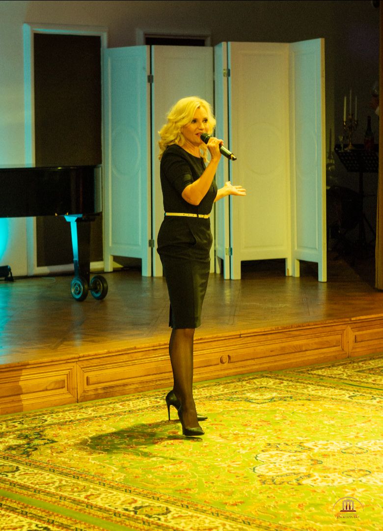 Egy csinos fekete ruhás nő áll a képen, a kezében mikrofon van és éppen valamit énekel