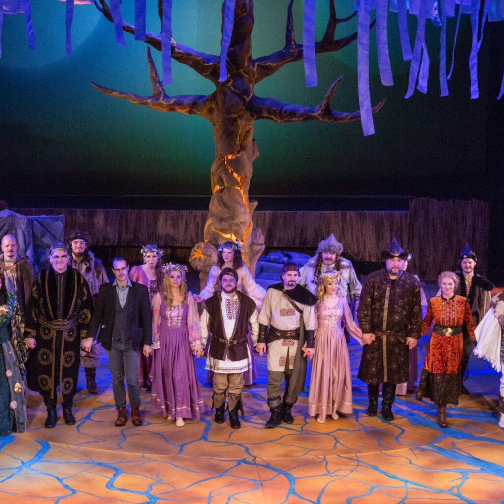 A Rege a csodaszarvasról összes szereplője a színpadon áll, fogják egymás kezét és mosolyognak a közönségre. A háttérben egy díszlet fa áll, aminek a textil csík levelei belógnak fentről a képbe