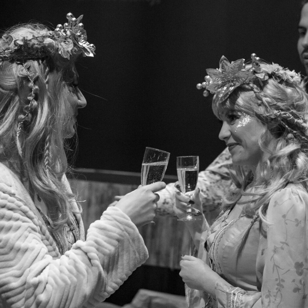 A fekete-fehér képen két tündérnek öltözött nő áll, akik éppen pezsgőspohárral koccintanak.