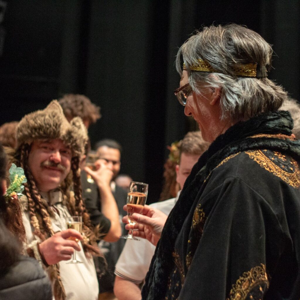 EGy fekete palástos ruhát és vékony koronát viselő férfi áll a képen, kezében pezsgős pohár van. A háttérben egy ősmagyar ruhát viselő férfi beszélget több emberrel