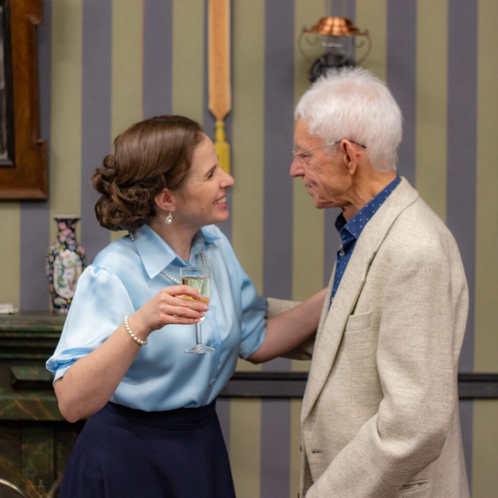 Egy nő és egy férfi áll egymással szemben, mosolyognak egymásra. A nő kezében egy pezsgővel töltött pohár van