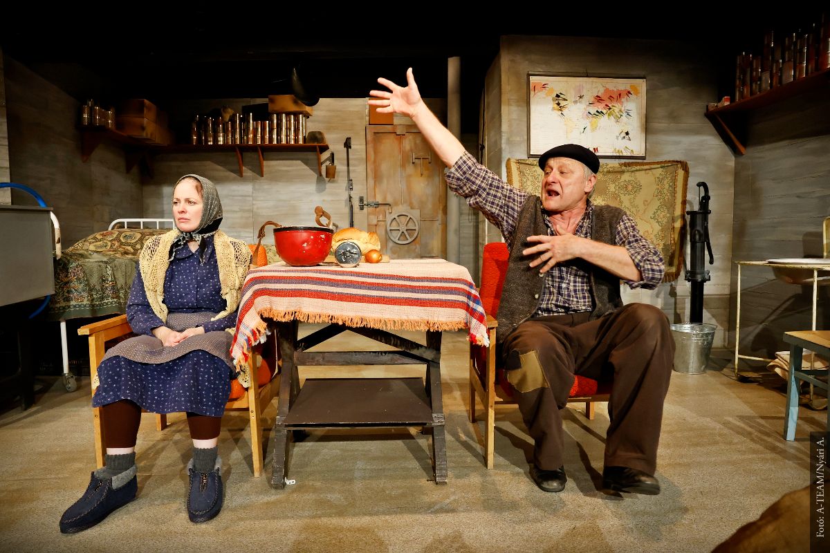 A színművészek a Nyolcvan vödör levegő című előadásban jelenet közben a színpadon, asztal körül ülnek.